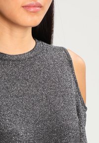 Kvinna som bär en ärmlös, glittrig silverblus med hög krage, delvis vy från axlarna till läpparna mot en enkel ljus bakgrund.