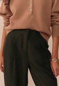 Pulover bej tricotat cu trei nasturi la guler; pantaloni drepți verde închis, cu buzunare laterale; textură netedă și croială lejeră.