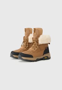 Brune isolerte vinterstøvler med en fluffy mansjett, svart gummitå og Vibram såle, plassert mot en enkel hvit bakgrunn.