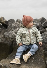 Vadderad grön jacka med blommönster, grå fleecefickor, blå jeans och bruna skor med kardborre. Barn sitter på stenar.