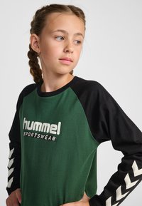 Maglietta a maniche lunghe verde e nera con texture a coste, con logo bianco "hummel SPORTSWEAR" e accenti a zigzag sulle maniche.