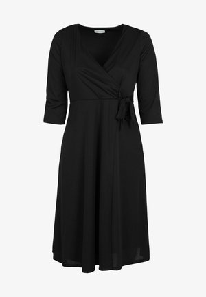 Kvinde iført en sort, knælange kjole med trekvartærmer og røde højhælede støvler, stående med ryggen mod beskueren.