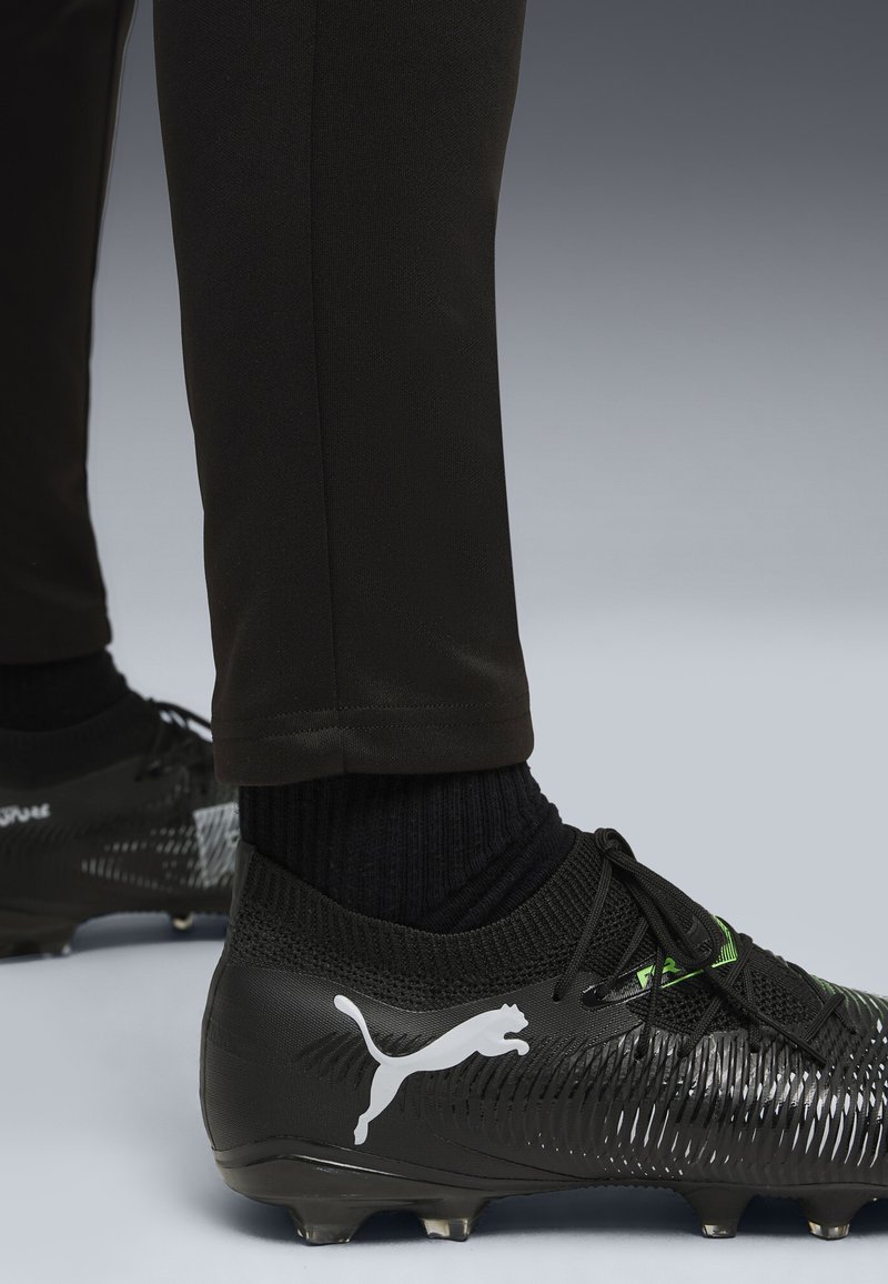 Svart fotbollssko med texturerad syntetisk ovandel, vit logotyp och gröna detaljer. Svarta strumpor synliga ovanför åtsittande svarta byxor.