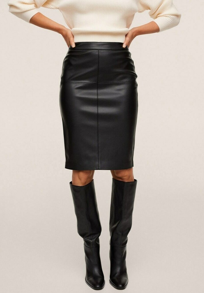 Femme portant une jupe crayon en cuir noir et des bottes en cuir noires montant jusqu'aux genoux, debout les mains sur les hanches devant un fond uni.