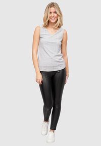 Grå ermeløs topp med blonderdesign, kombinert med svarte faux lær leggings og hvite sneakers. Modellen står på en nøytral bakgrunn.
