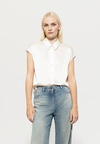 Weißes Seidenhemd mit spitzem Kragen und kurzen, hochgerollten Ärmeln, kombiniert mit hellblauen, weit geschnittenen Jeans, die eine ausgebleichte Textur aufweisen.