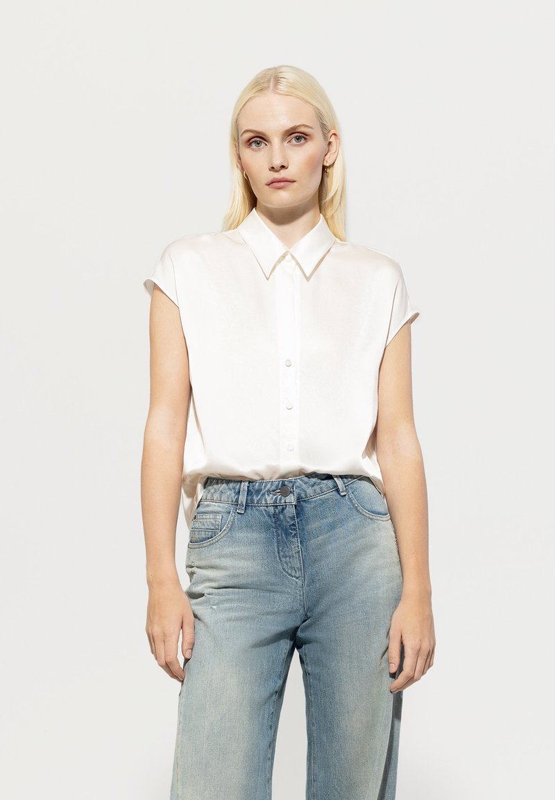 Weißes Seidenhemd mit spitzem Kragen und kurzen, hochgerollten Ärmeln, kombiniert mit hellblauen, weit geschnittenen Jeans, die eine ausgebleichte Textur aufweisen.