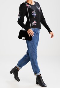 Un pull noir avec broderies florales, un jean en denim et des bottines à talons noires. Les accessoires comprennent un petit sac à main noir avec une bandoulière en chaîne.