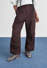 Pantaloni cargo marroni con vestibilità relax, dotati di tasche profonde, vita con cordoncino e dettagli di cucitura sottili. Abbinati a scarpe da trekking grigie.