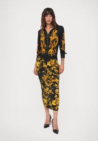 Blusa preta e saia com padrão barroco amarelo, corte justo, mangas longas, e design de saia lápis, combinadas com sapatos de salto alto.