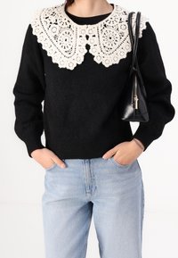 Pull noir avec un grand col en crochet crème, poignets et ourlet côtelés, associé à un jean bleu clair et un petit sac à main noir.