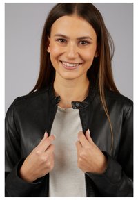 Black leather jacket with a rounded collar, silver zipper, and smooth texture, worn over a light gray shirt. Model holds jacket closed.