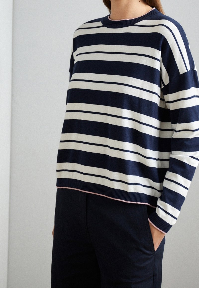 Personne portant un pull à manches longues bleu marine et blanc à rayures horizontales, les mains dans les poches d’un pantalon foncé.
