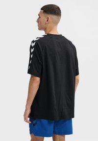 Zwarte t-shirt met korte mouwen en witte chevronpatroon op de mouwen, met een ontspannen fit en een gladde textuur. Draagt met blauwe shorts.