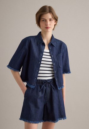 Femme portant une chemise en jean bleu foncé courte boutonnée et un short assorti avec des bords effilochés, associée à un débardeur rayé blanc et noir.