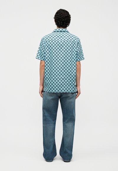 Άνδρας με σγουρά μαλλιά που φοράει μπλε και μαύρο καρό κοντομάνικο πουκάμισο και φαρδιά μπλε τζιν, στεκόμενος με την πλάτη σε απλό λευκό φόντο.