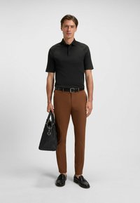 Svart kortärmad polo, bruna slim-fit byxor, svarta loafers och en strukturerad svart tote-väska på en neutral bakgrund.