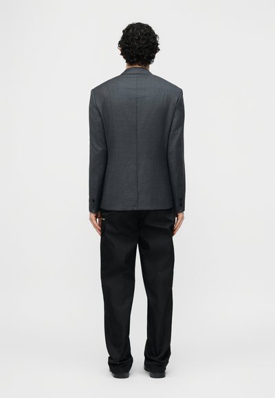 Homme aux cheveux bouclés vu de dos, portant un blazer gris et un pantalon noir ample, sur un fond blanc uni.