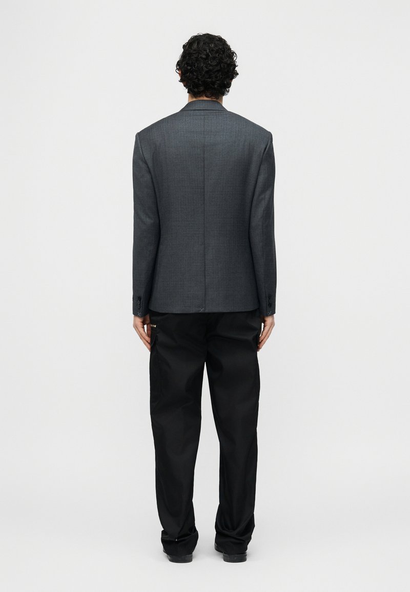 Homme aux cheveux bouclés vu de dos, portant un blazer gris et un pantalon noir ample, sur un fond blanc uni.