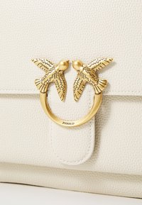 Borsa a mano in pelle beige chiaro con hardware circolare dorato decorato da due uccelli e superficie texturizzata. Logo visibile.