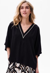 Schwarzes, locker sitzendes Oberteil mit V-Ausschnitt und kurzen Ärmeln, mit einem dekorativen silbernen Saum entlang des Ausschnitts. Glatte Stoffstruktur.