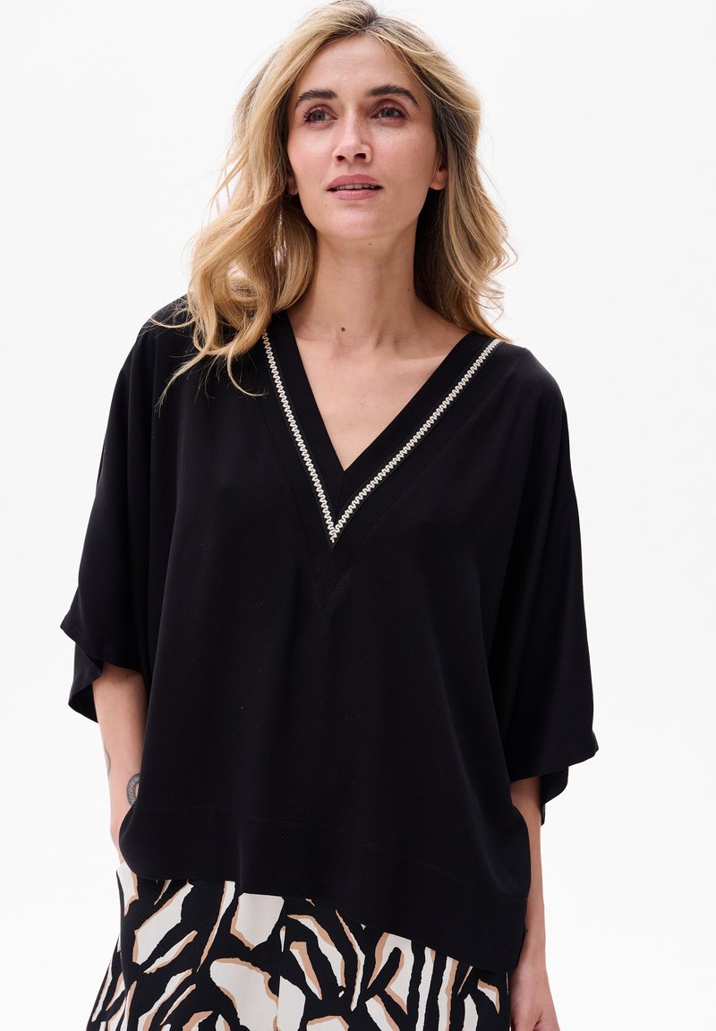 Schwarzes, locker sitzendes Oberteil mit V-Ausschnitt und kurzen Ärmeln, mit einem dekorativen silbernen Saum entlang des Ausschnitts. Glatte Stoffstruktur.