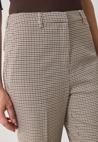 Gros plan sur une personne portant un pantalon à carreaux beige et marron et une chemise marron, la main gauche relaxée sur le côté.