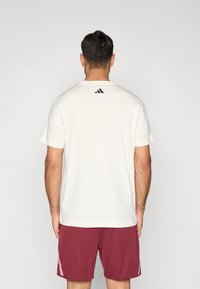 Men's white T-shirt with short sleeves, featuring a small black logo on the back, paired with maroon shorts. Smooth texture and basic design.