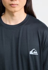 Černé tričko s kulatým výstřihem a krátkými rukávy. Obsahuje bílé logo na levé části hrudi. Hladká textura látky.
