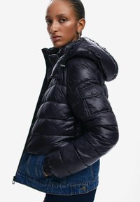 Giacca nera imbottita con finitura lucida, dotata di cappuccio, chiusura con zip e una sezione inferiore in denim con tasche e dettagli cuciti.