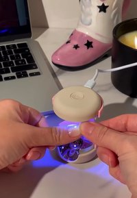 Nagellamp met een ronde beige bovenkant, roze accenten, die heldere, verzorgde nagels verlicht. Een laptop en een zwarte kaars staan op de achtergrond.