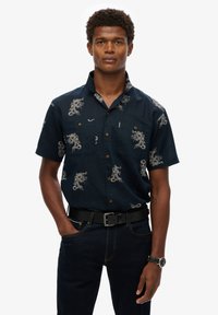 Camisa de manga corta azul marino con patrones de dragones blancos, dos bolsillos en el pecho con botones y un cuello. Llevada con jeans oscuros y un cinturón negro.