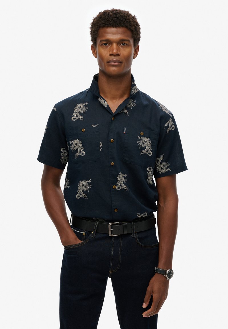 Camisa de manga corta azul marino con patrones de dragones blancos, dos bolsillos en el pecho con botones y un cuello. Llevada con jeans oscuros y un cinturón negro.