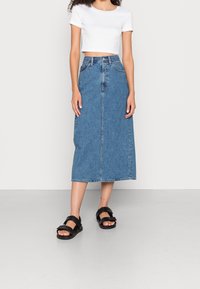 Jeanskjol i medelblått, högmidjad, knä-lång med rak passform; matchad med en vit kortärmad t-shirt och svarta sandaler.