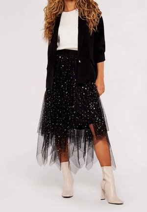 Vrouw draagt een zwarte tule rok met sterpatroon, een witte geribbelde top, een zwart colbert en witte enkellaarsjes met hoge hakken tegen een effen achtergrond.