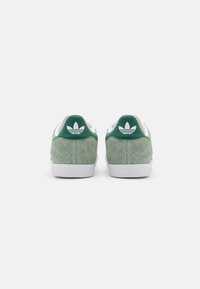 Gröna mockasneakers med vita gummisulor, med en grön hälflik och vit logotypaccent. Texturerat material och enkel design.