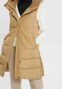 Gilet imbottito beige con cappuccio, dotato di zip frontale, dettagli a coste e tasche laterali. Design trapuntato con taglio alla lunghezza del ginocchio.