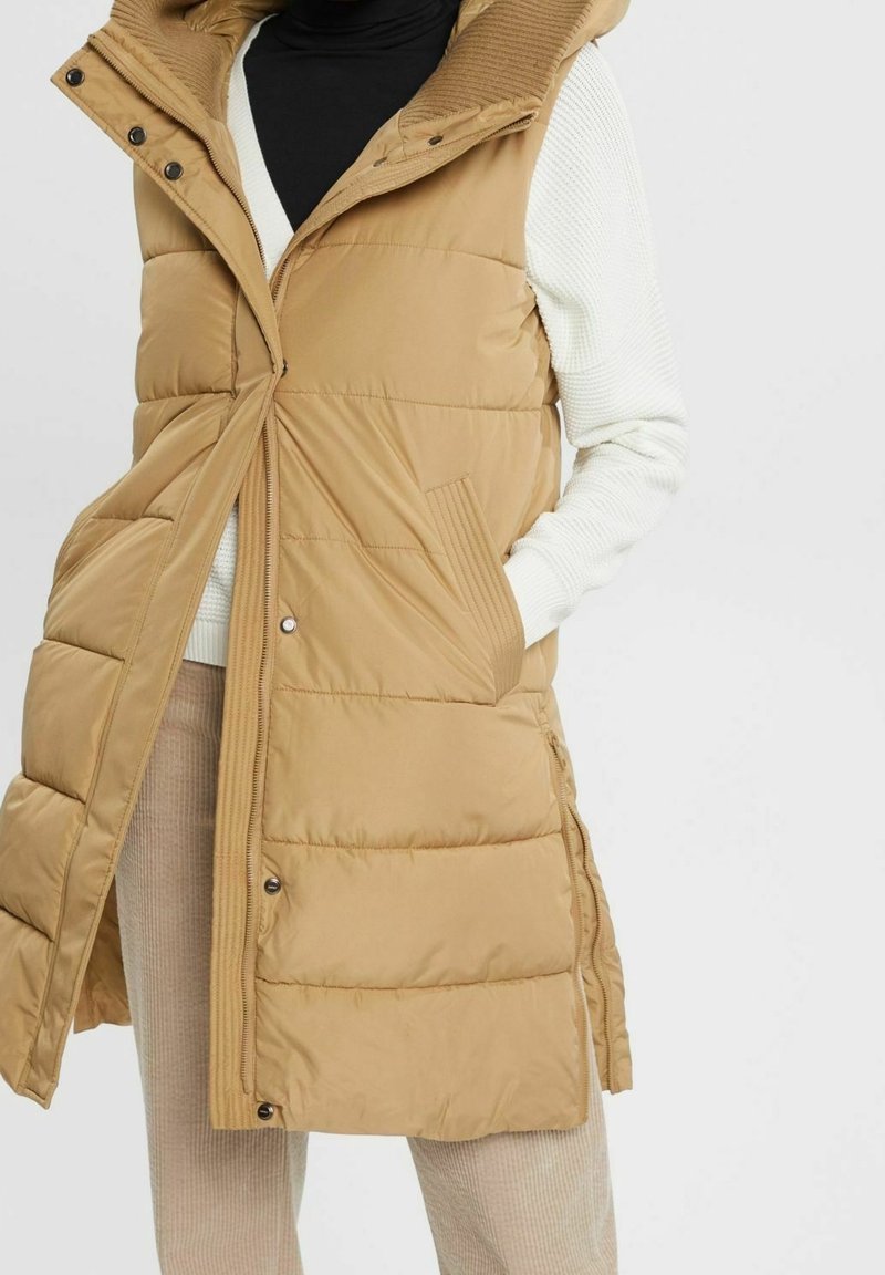 Gilet imbottito beige con cappuccio, dotato di zip frontale, dettagli a coste e tasche laterali. Design trapuntato con taglio alla lunghezza del ginocchio.