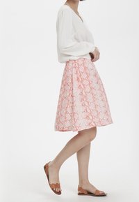 Una gonna al ginocchio di un delicato colore rosa chiaro con un intricato motivo di pelle di serpente bianca, abbinata a sandali piatti marroni e a una blusa bianca.