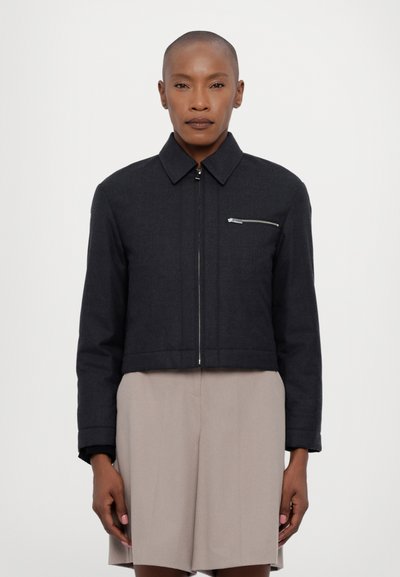 Veste noire cintrée avec un col structuré, fermeture éclair argentée et poche zippée sur la poitrine. Tissu lisse avec un design épuré.