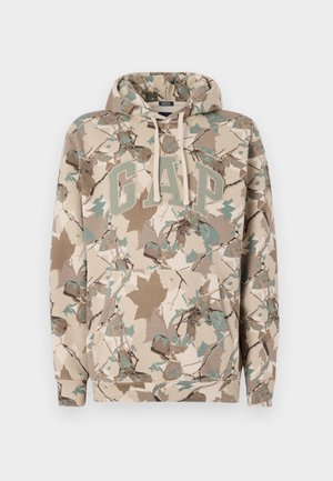 Sudadera beige con un patrón de hojas de camuflaje, que presenta un gran logo "GAP" en verde y un bolsillo frontal. Fabricada con un tejido suave y texturizado.