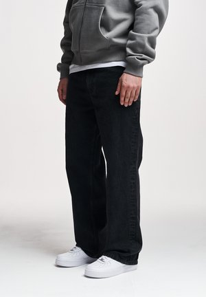 Oseba z oblečeno sivo kapucarno jakno z zadrgo, črnimi širokimi kavbojkami in belimi supergami, stoji pred enobarvnim svetlim ozadjem.