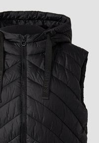 Schwarze gesteppte ärmellose Pufferweste mit Kapuze, Frontreißverschluss und schwarzen Zugbändern mit der Aufschrift "S.Oliver" vor einem schlichten hellen Hintergrund.