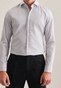 Uomo che indossa una camicia a righe verticali bianche e grigie infilata in pantaloni neri su misura, in piedi contro uno sfondo neutro.