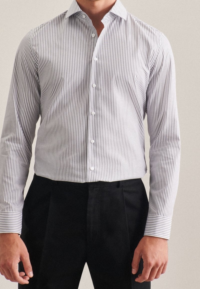 Uomo che indossa una camicia a righe verticali bianche e grigie infilata in pantaloni neri su misura, in piedi contro uno sfondo neutro.