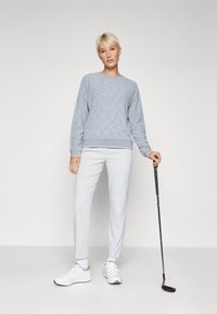 Ljusgrå quiltad sweatshirt med rund halsringning och ribbade ärmslut, parat med ljusgrå slim-fit byxor och vita sneakers, som håller i en golfklubba.