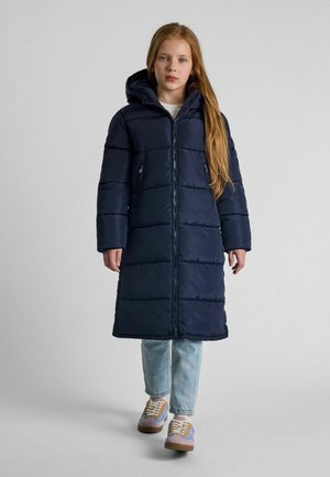 Giovane che indossa un lungo cappotto imbottito blu navy con cappuccio, jeans blu chiaro e sneakers multicolori, in piedi contro uno sfondo neutro.