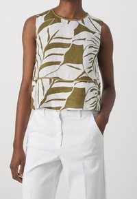 Top court sans manches en lin blanc avec des motifs de feuilles vertes. Encolure arrondie, double couche, porté avec un pantalon blanc.