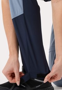 Persoon die de klittenbandsluiting aan de onderkant van het donkerblauwe broekspijpje aanpast, over een zwarte waterdichte wandelschoen.