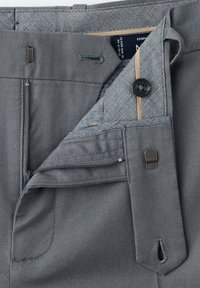 Taille de pantalon gris montrant une fermeture éclair partiellement ouverte, un bouton et une fermeture à pince métallique, avec une étiquette intérieure visible et des détails de couture.